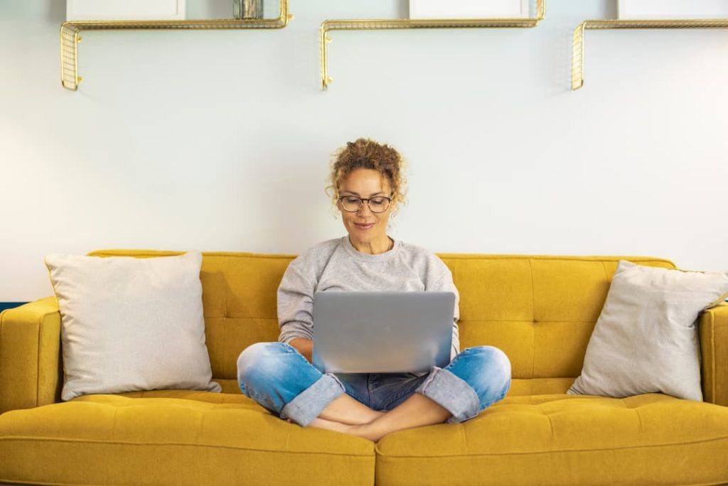 One woman smiling and using laptop computer at home sitting comfortably on a yellow sofa in living room.